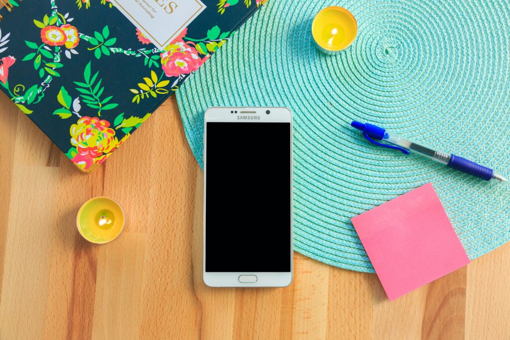 An image of a cell phone with a blue placemat, post its, a pen, and a notebook.